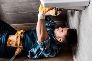 Plumber in blue shirt and gloves working under a bathroom sink, highlighting professional installation of eco-friendly showerheads to prevent leaks and ensure energy efficiency.
