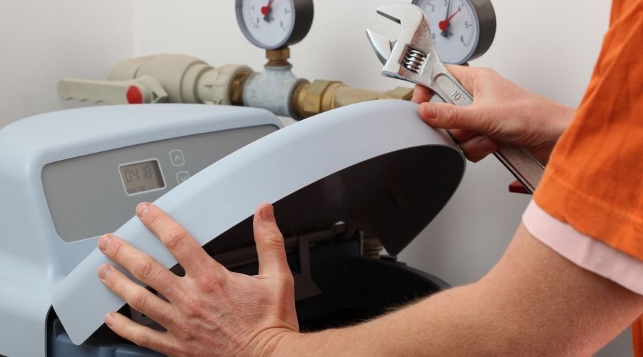 Person using a wrench to open a water softener, with pressure gauges and plumbing components visible, illustrating maintenance of plumbing systems to prevent hard water issues.