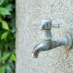 Metal faucet on textured gray stone wall with green foliage; tips for winter plumbing preparation by Lodder Brothers Limited.