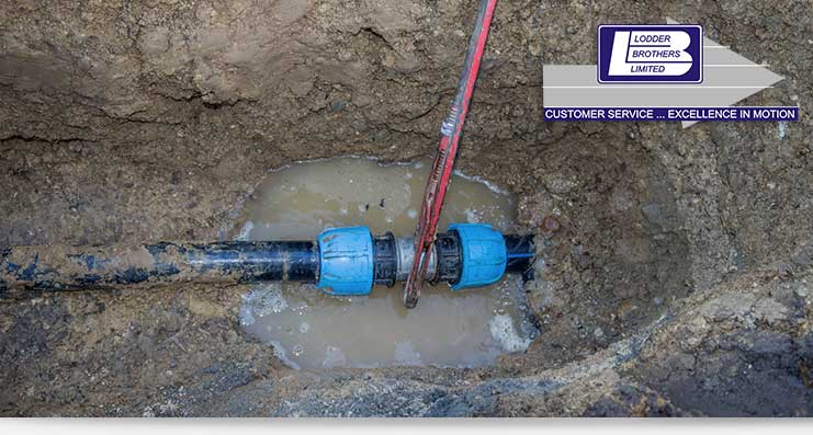 Commercial burst pipe repair in Guelph, featuring a plumbing technician using a wrench on a blue pipe connector in a muddy trench, with Lodder Brothers Limited logo visible.