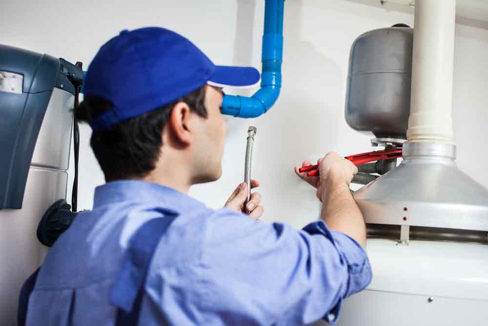 Plumber adjusting a gas water heater pilot light with a wrench, surrounded by plumbing components and pipes.